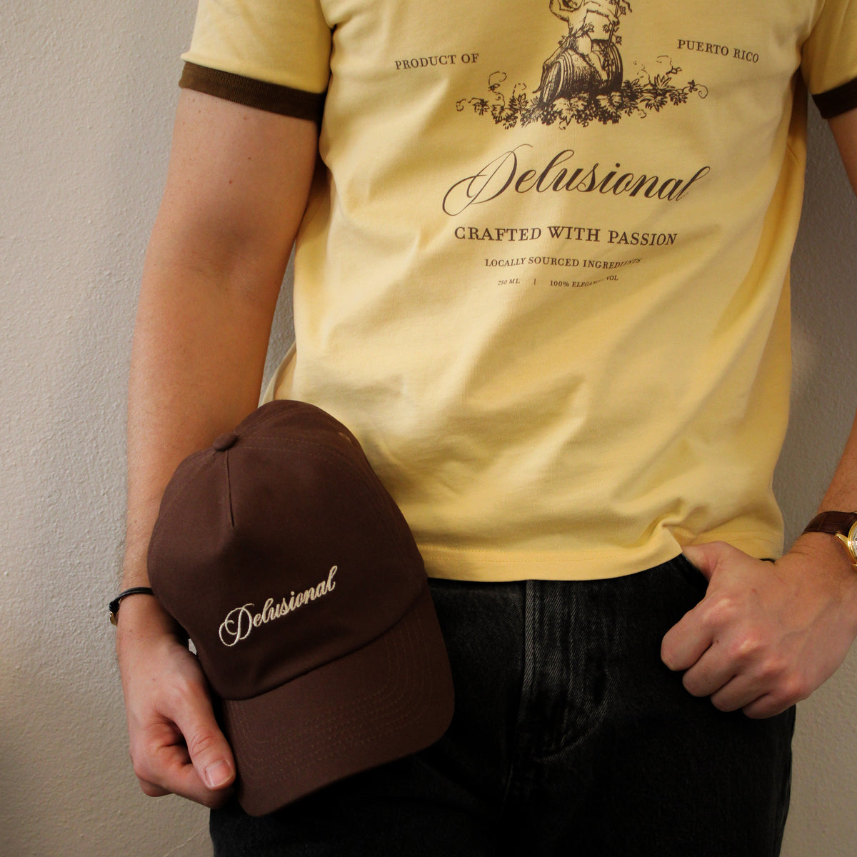 A man holding a brown cap with 'delusional' embroidered in white color, wearing a pale yellow graphic shirt.