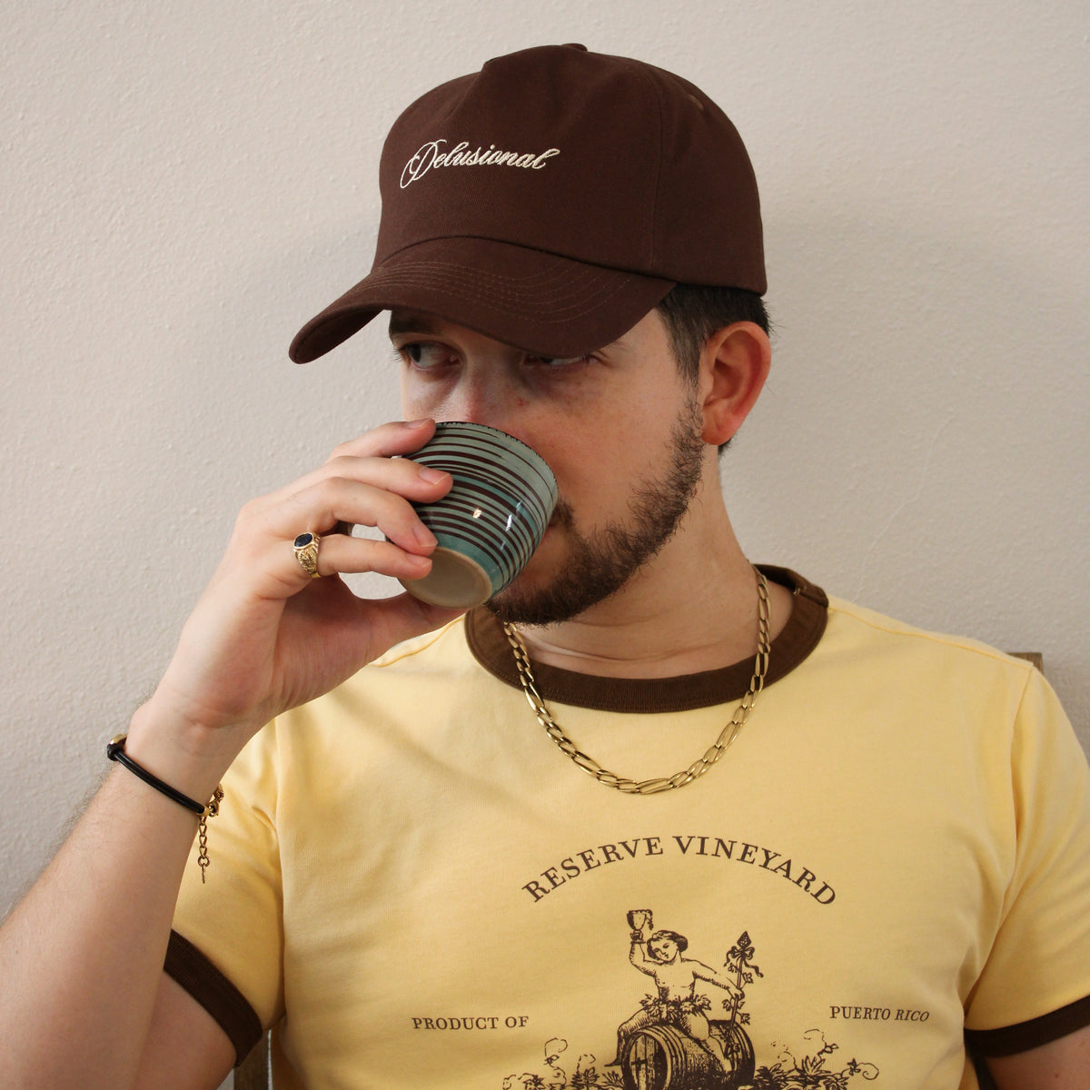A man wearing a brown cap drinking a cup of coffee with a pale yellow ringer T-shirt with brown rims.