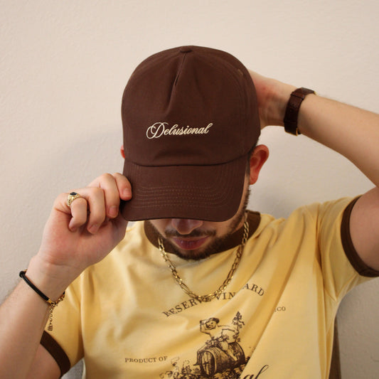 A man wearing a dark brown baseball cap with 'Delusional' embroidered in white script on the front, holding the brim over his eyes. He is wearing a yellow graphic t-shirt and a gold chain necklace against a plain beige wall.