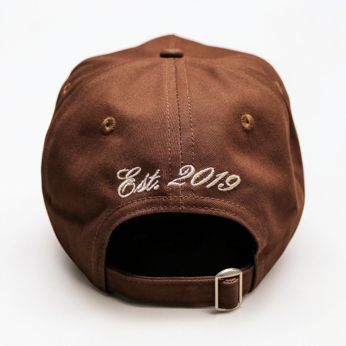 A brown canvas baseball cap shown from the back, featuring 'Est. 2019' embroidered in white script on the back panel and an adjustable strap with a silver metal buckle.