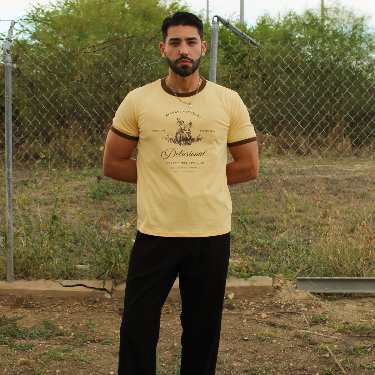 A man wearing a pale yellow graphic t-shirt with brown trim, black trousers, and black loafers, standing outdoors in front of a chain-link fence.