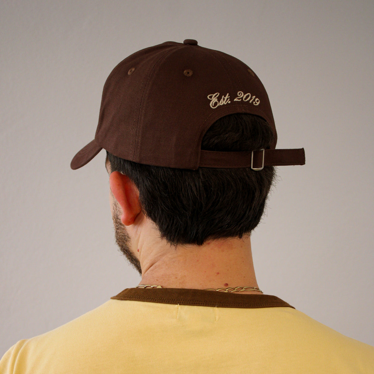A man viewed from behind wearing a dark brown baseball cap with 'Est. 2019' embroidered in beige script on the back, paired with a yellow t-shirt against a plain gray background.