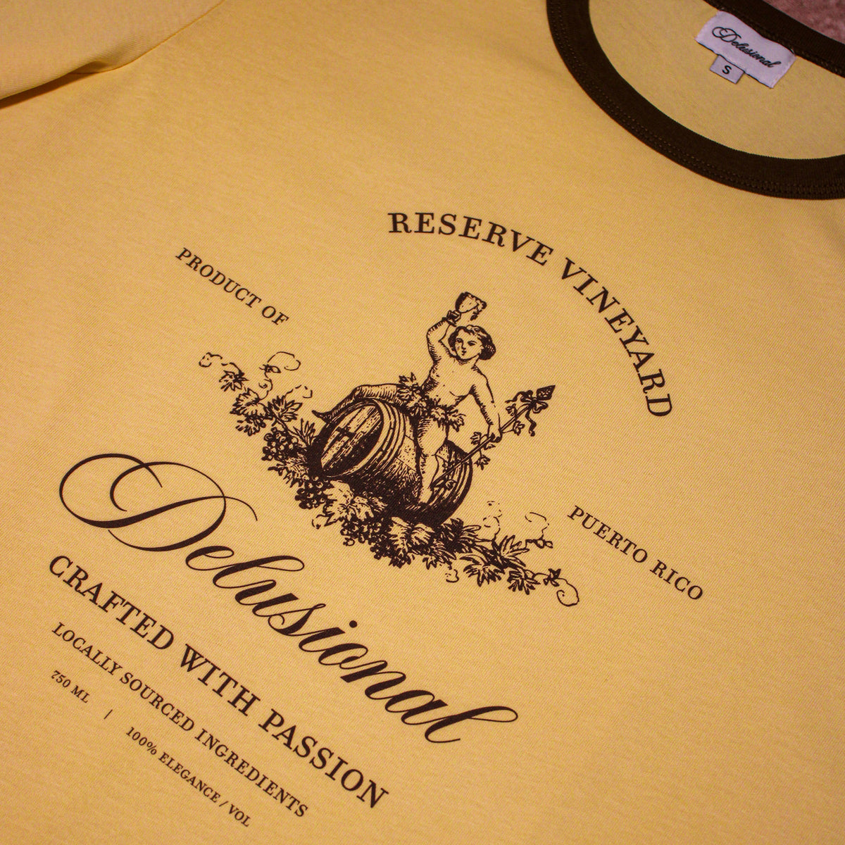 A close-up of a pale yellow t-shirt featuring a black graphic print with the text 'Reserve Vineyard' and 'Delusionál Crafted with Passion'. The shirt has a black collar trim and a visible neck label.