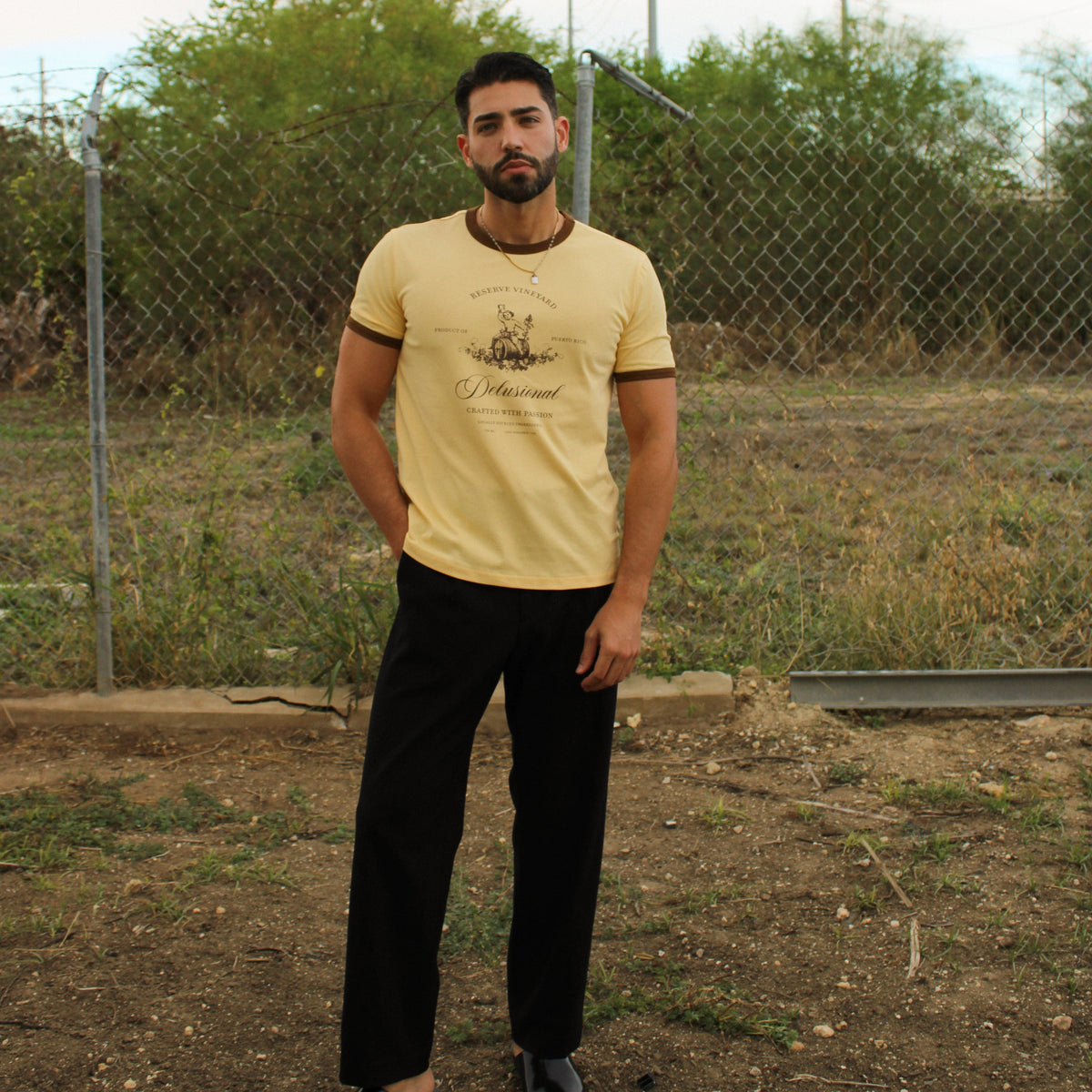 A man wearing a pale yellow graphic t-shirt with dark brown trim, black pants, and black loafers, standing outdoors in front of a chain-link fence.