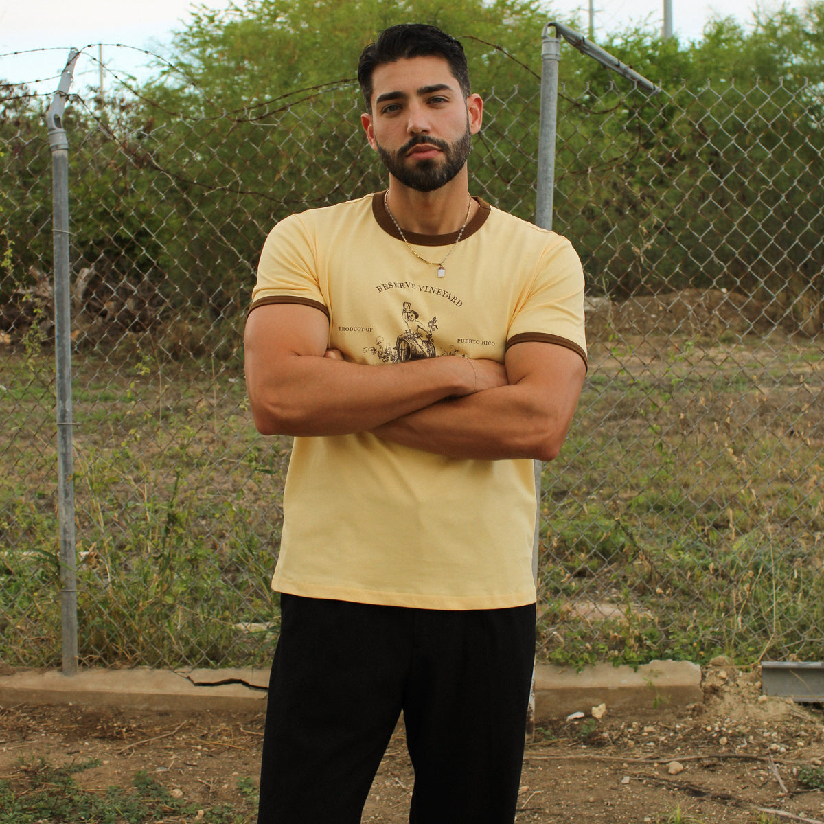 A man with a beard stands outdoors with his arms crossed, wearing a pale yellow graphic t-shirt and black pants, positioned in front of a chain-link fence and green bushes.