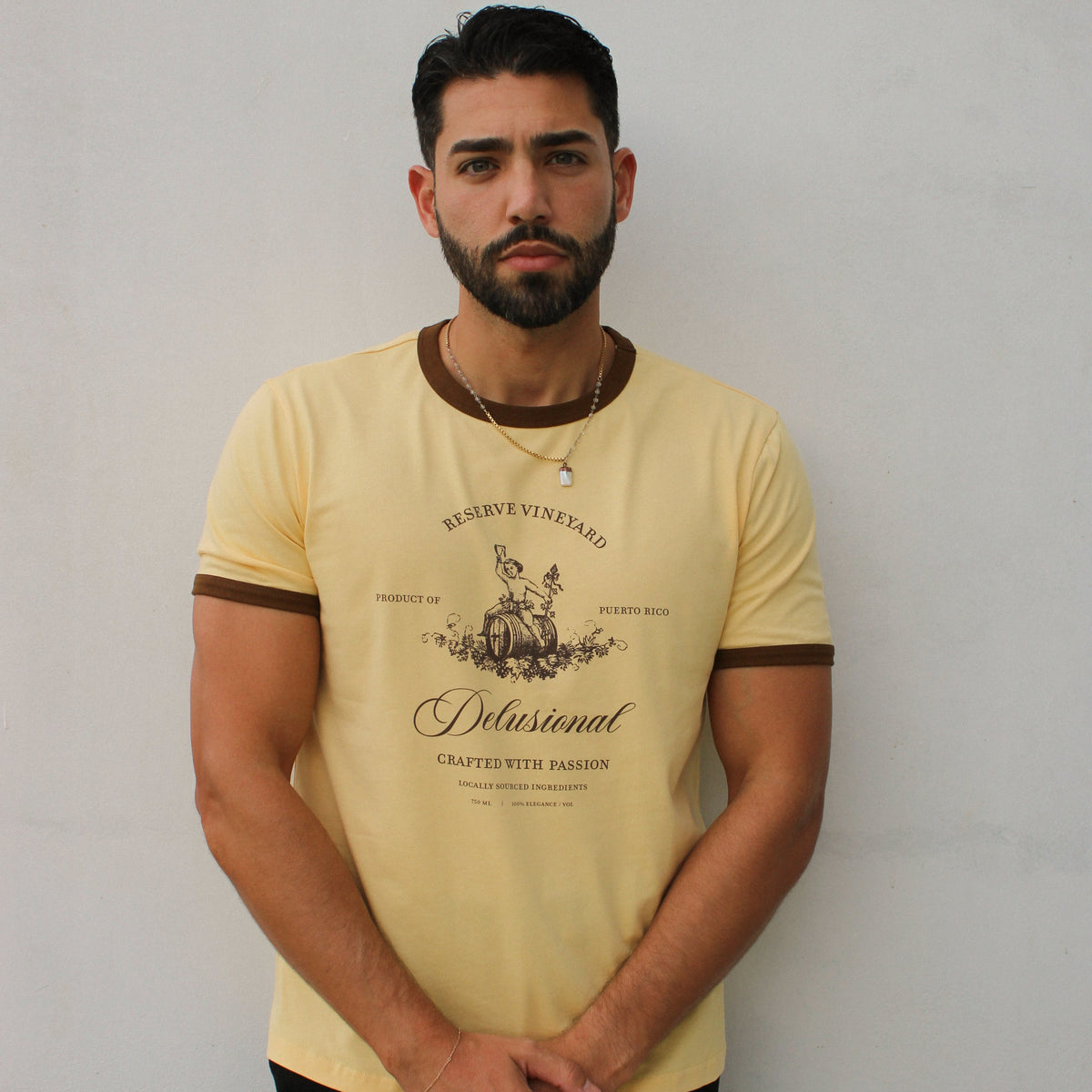 A man wearing a pale yellow ringer t-shirt with brown trim and a graphic print on the front, paired with black pants, standing against a plain light gray wall.