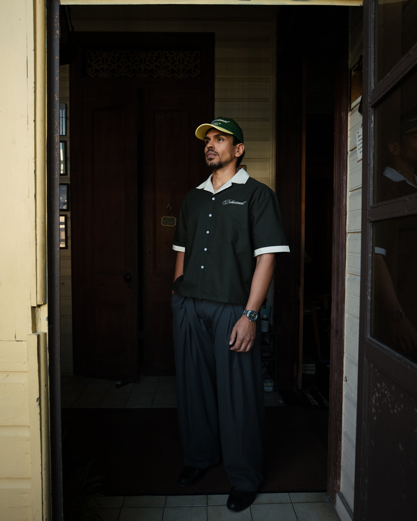 Man wearing designed in PR shirt with white trim and two-tone delusional hat, standing in a doorway.