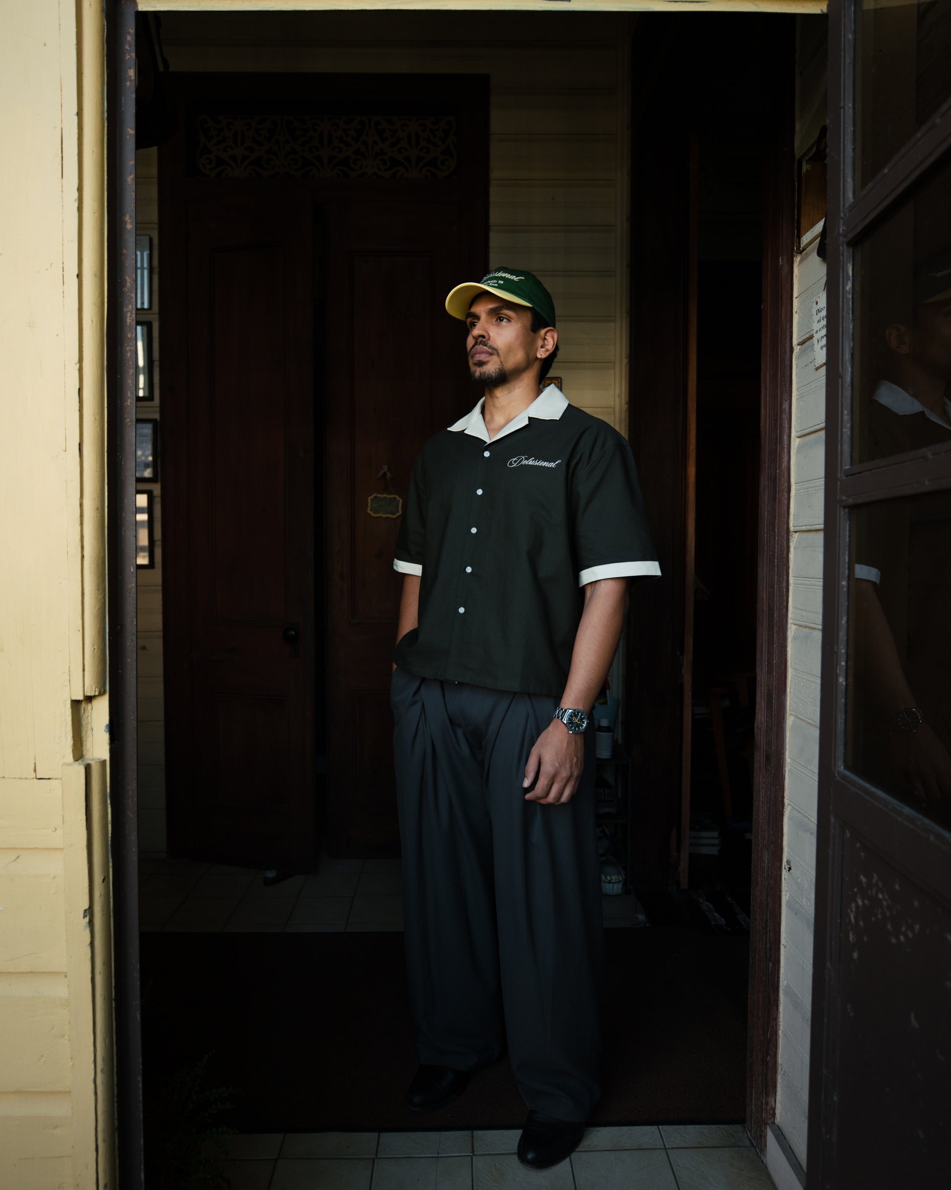 Man wearing designed in PR shirt with white trim and two-tone delusional hat, standing in a doorway.